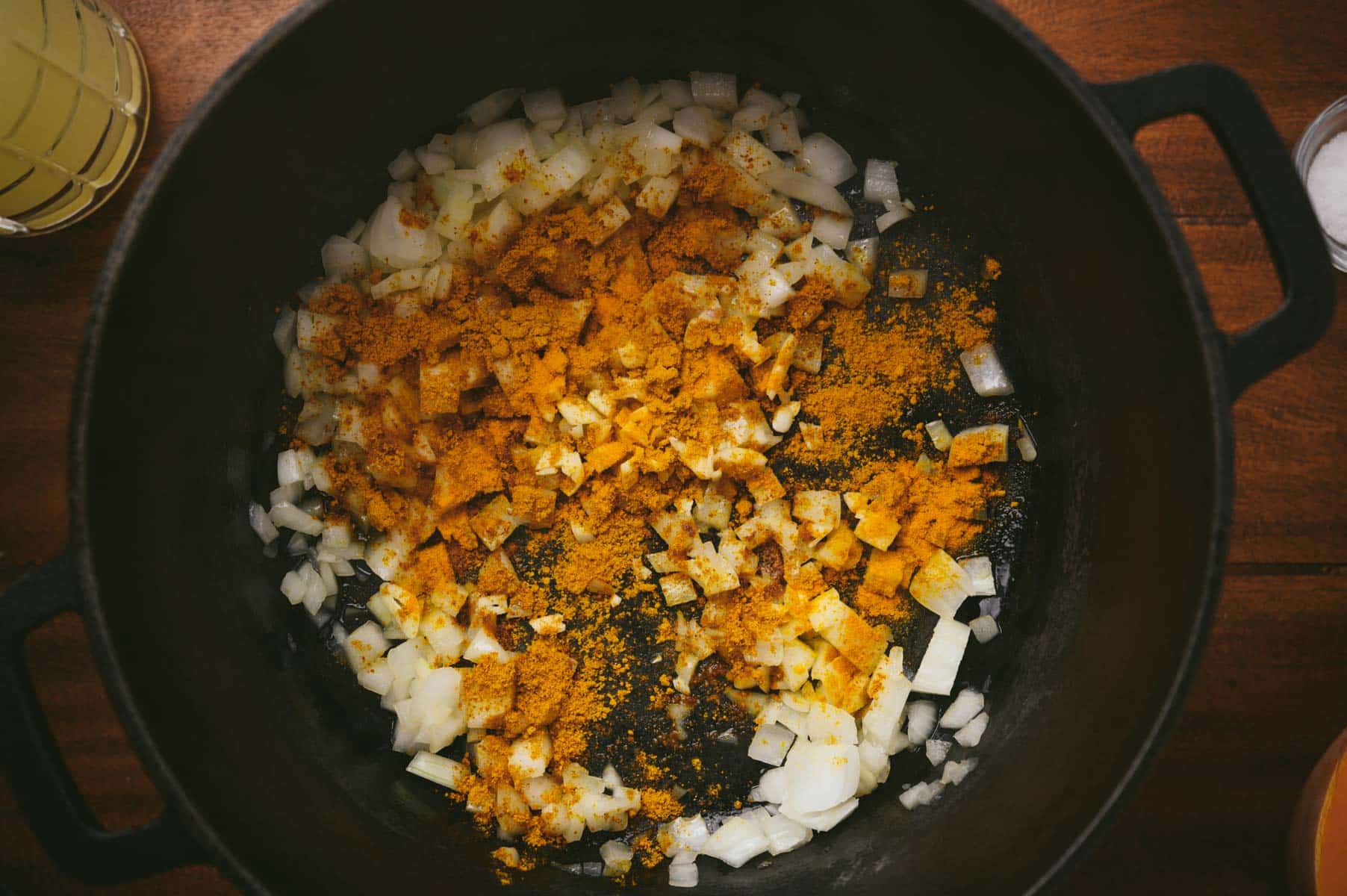Chopped onions and yellow spices cooking in a black pot on a wooden surface.