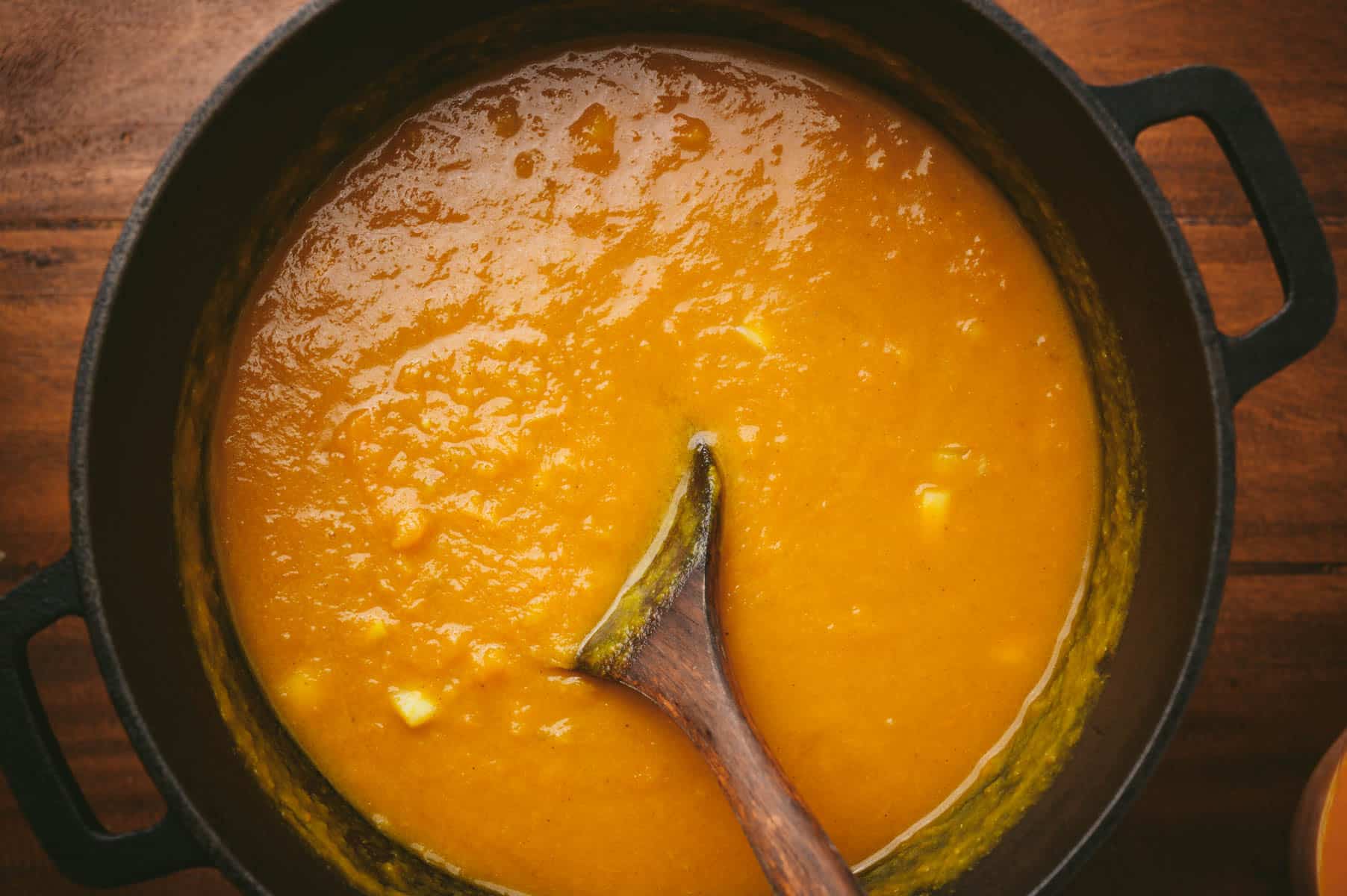 A wooden spoon in a pot of thick, orange pumpkin soup.