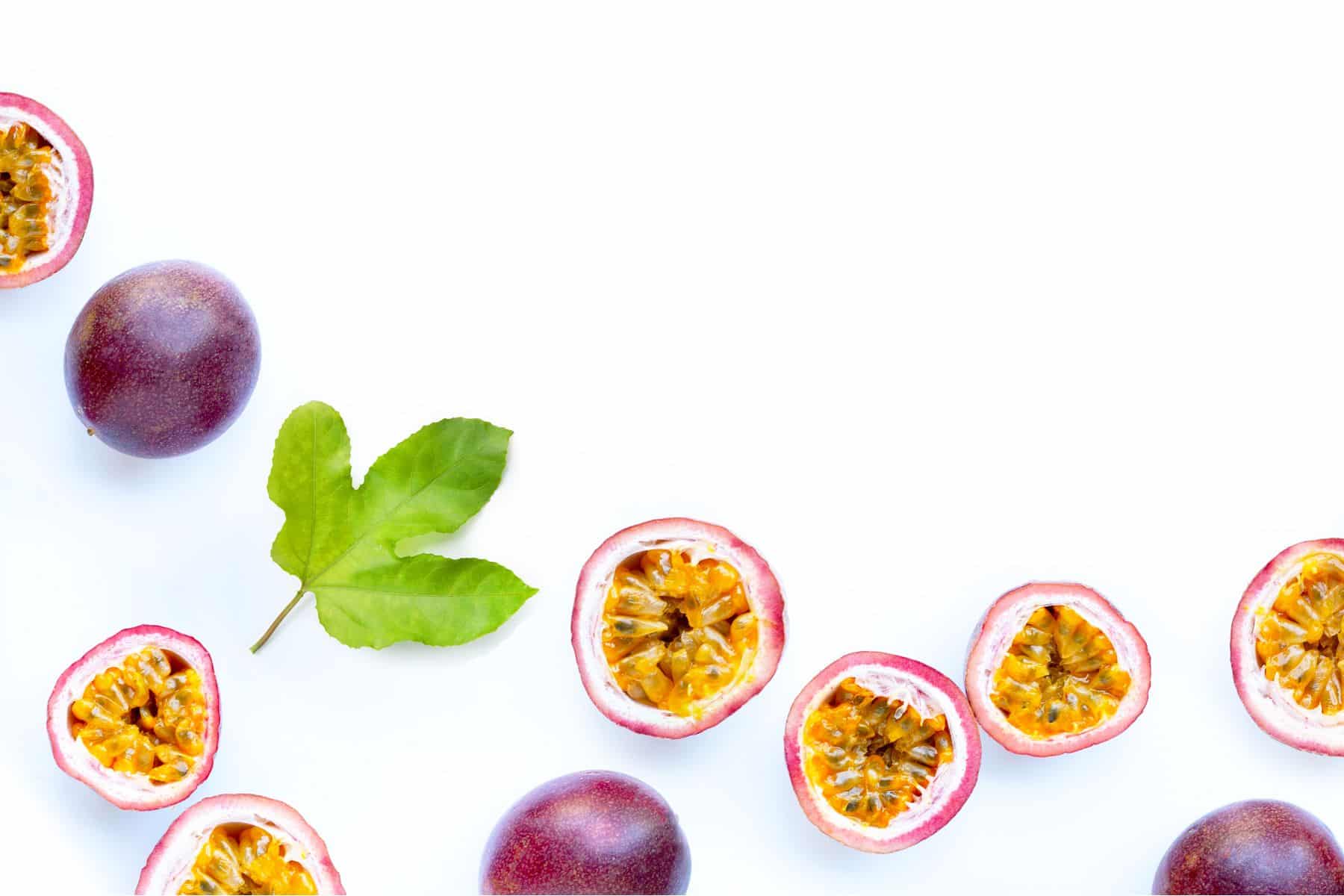 Purple passion fruits and a green leaf are arranged on a white background. Some fruits are halved, showing the yellow pulp and seeds.