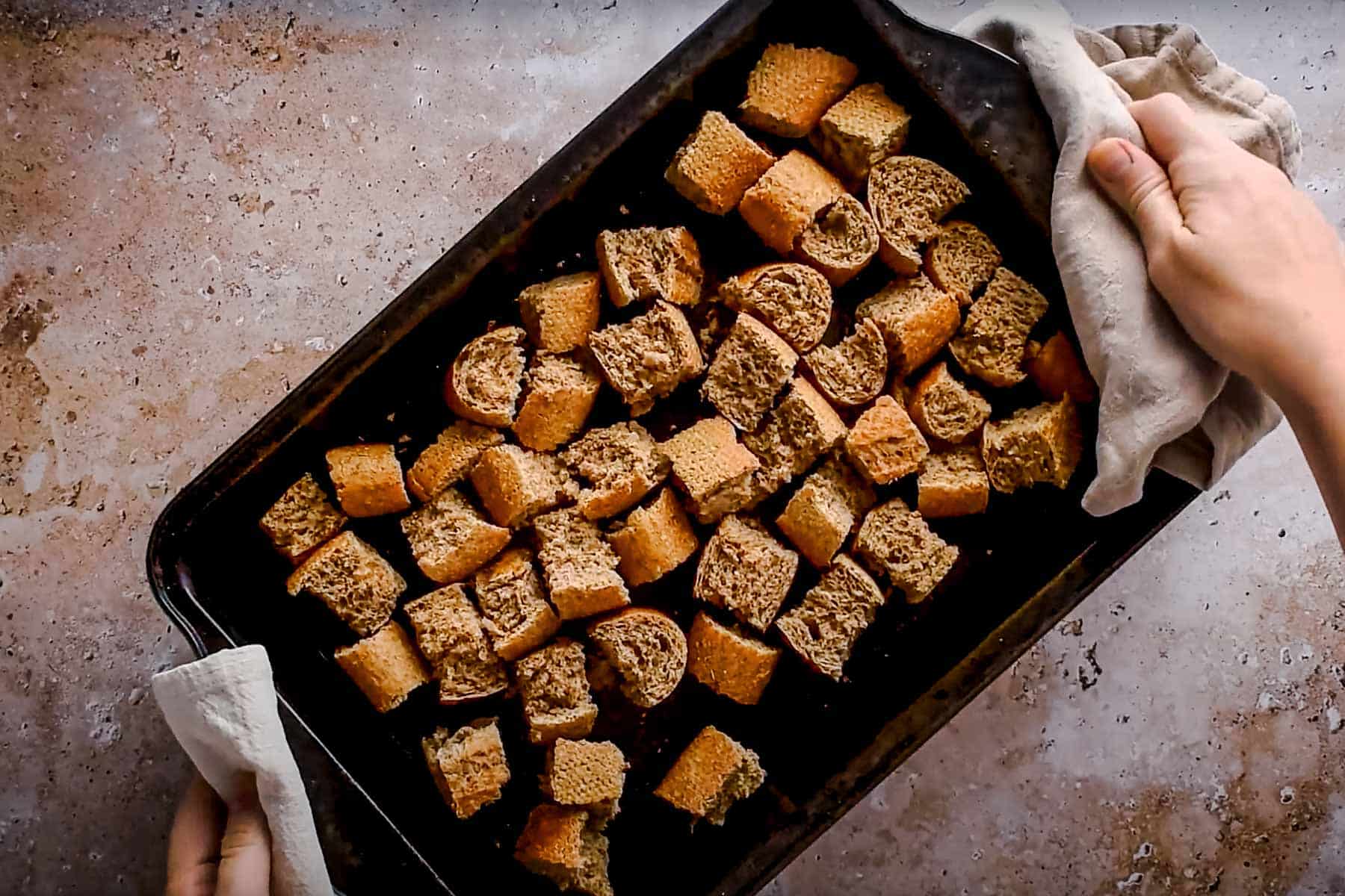 Hands holding a baking tray with toasted cubed bread pieces on a textured surface.