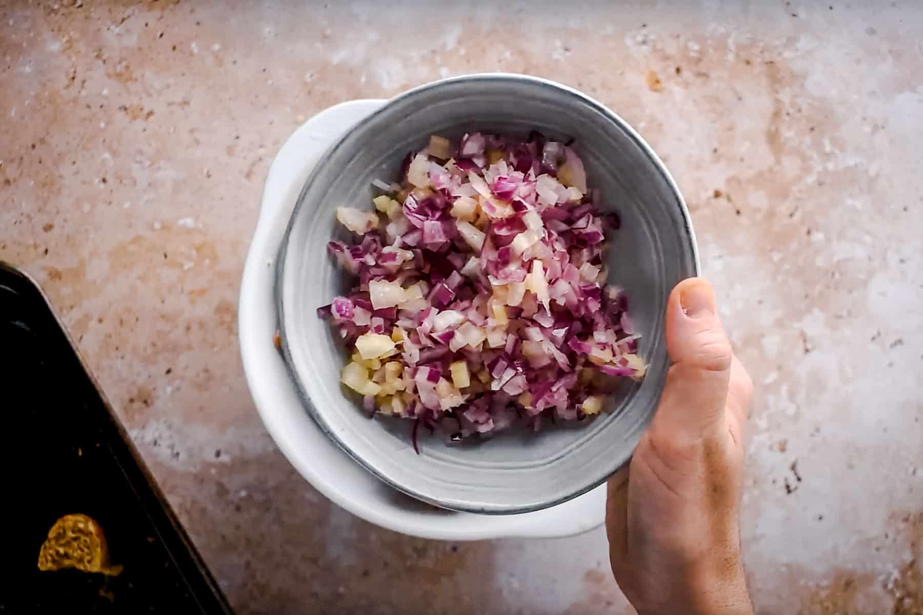 A bowl containing finely chopped red onions, held over a light-colored stone surface.
