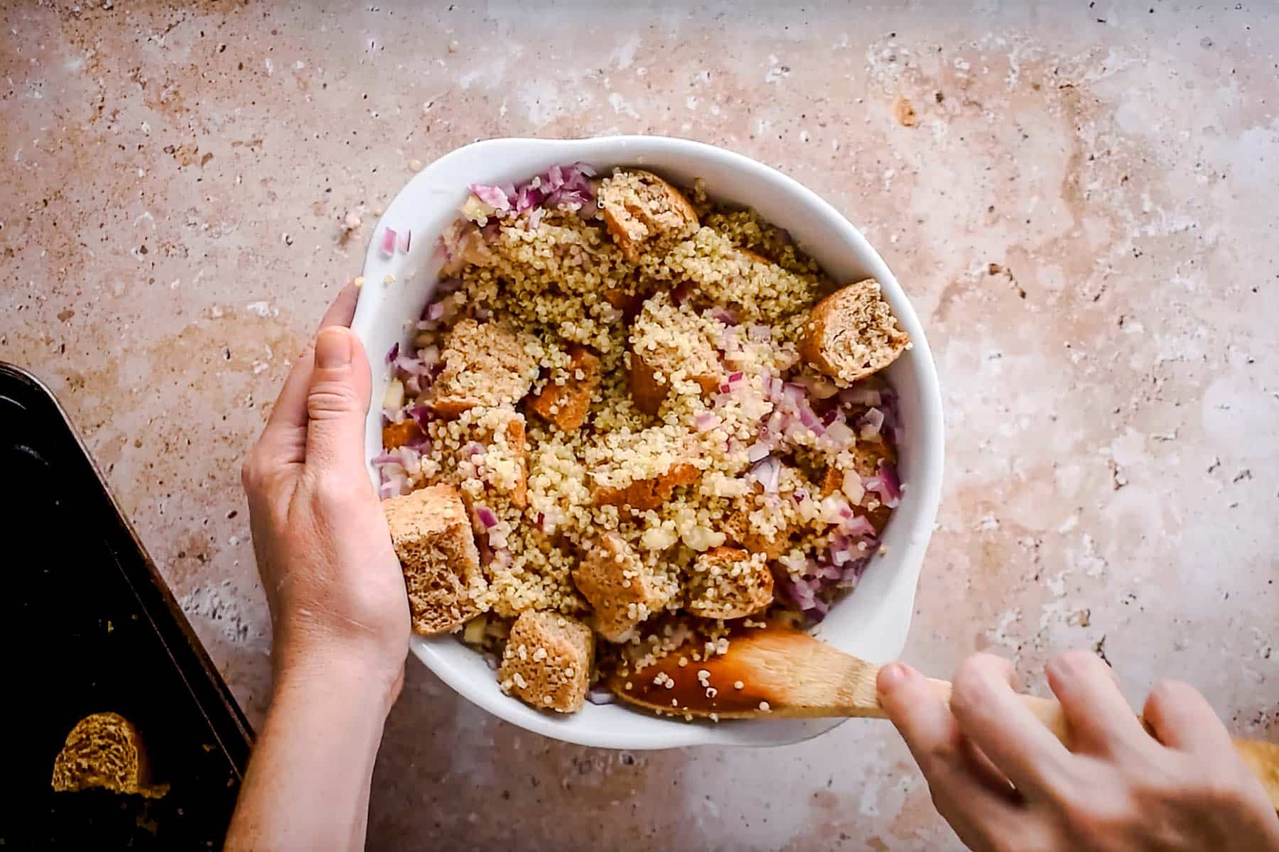 Hands mixing croutons, quinoa, and diced onions in a white bowl with a wooden spoon on a stone surface.