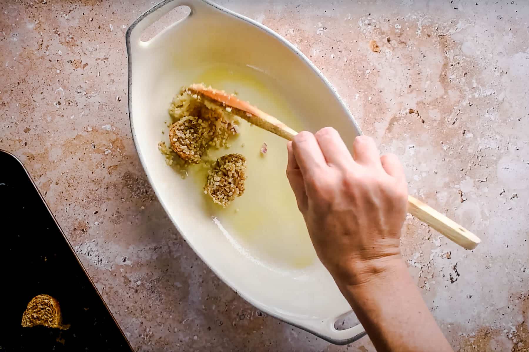 A hand using a wooden spoon to scoop stuffing into a white oval dish on a textured countertop.