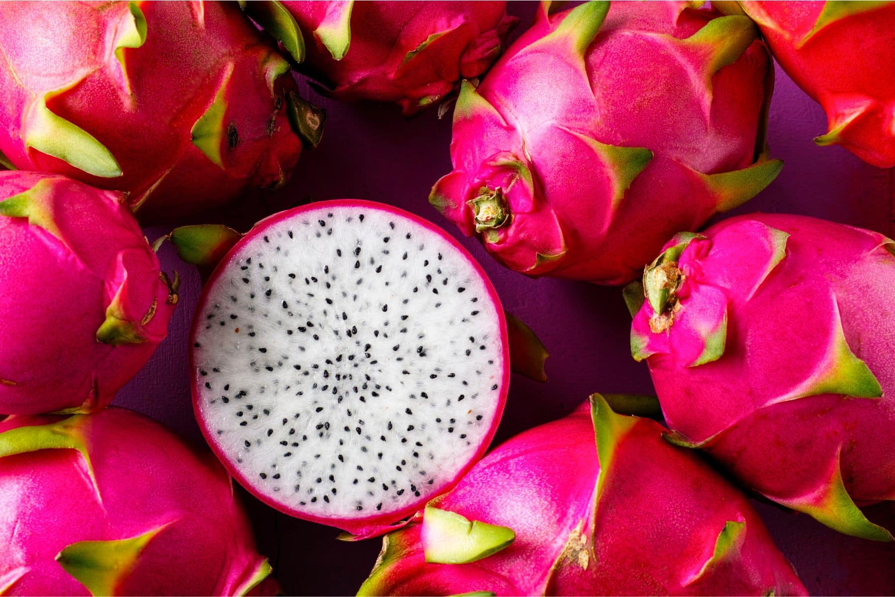 Pink dragon fruits with green tips surround a halved one, revealing white flesh with black seeds.