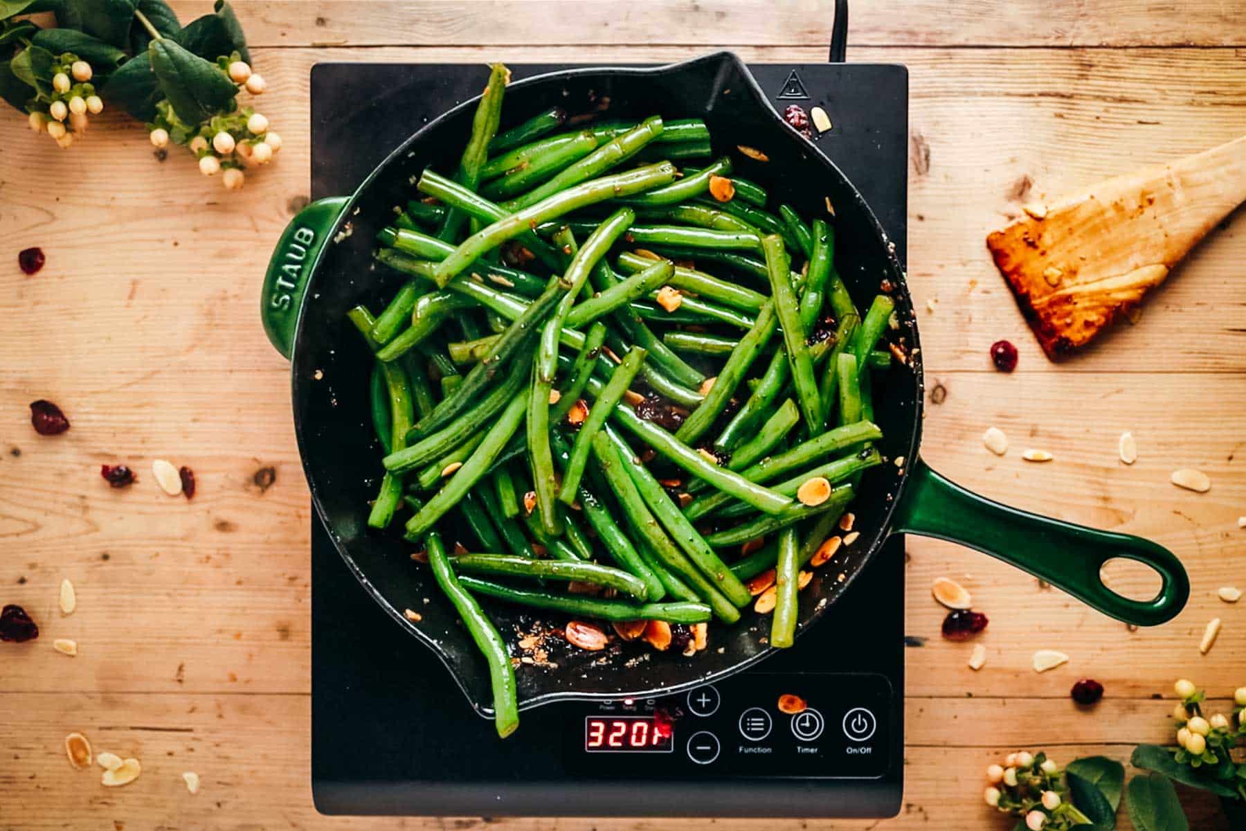 Sautéed green beans with almonds in a frying pan on an induction cooktop, surrounded by scattered nuts and berries.