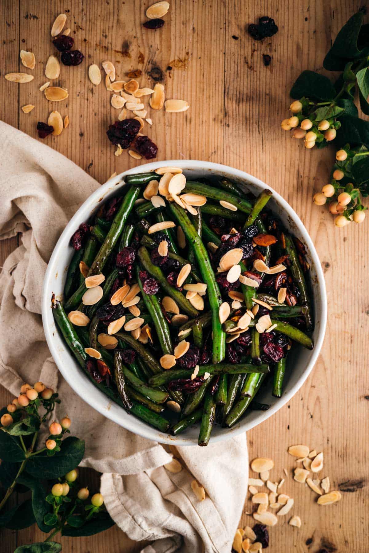 A bowl of green beans topped with sliced almonds and dried cranberries on a wooden table, surrounded by scattered nuts, berries, and foliage.