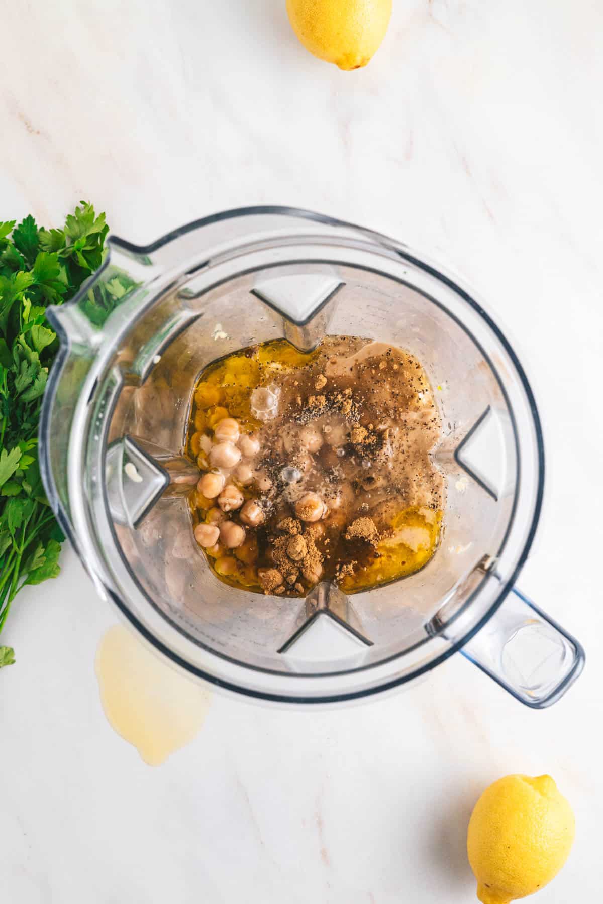 Top view of a blender containing chickpeas, spices, and liquid. Fresh parsley and whole lemons are nearby on a white surface.