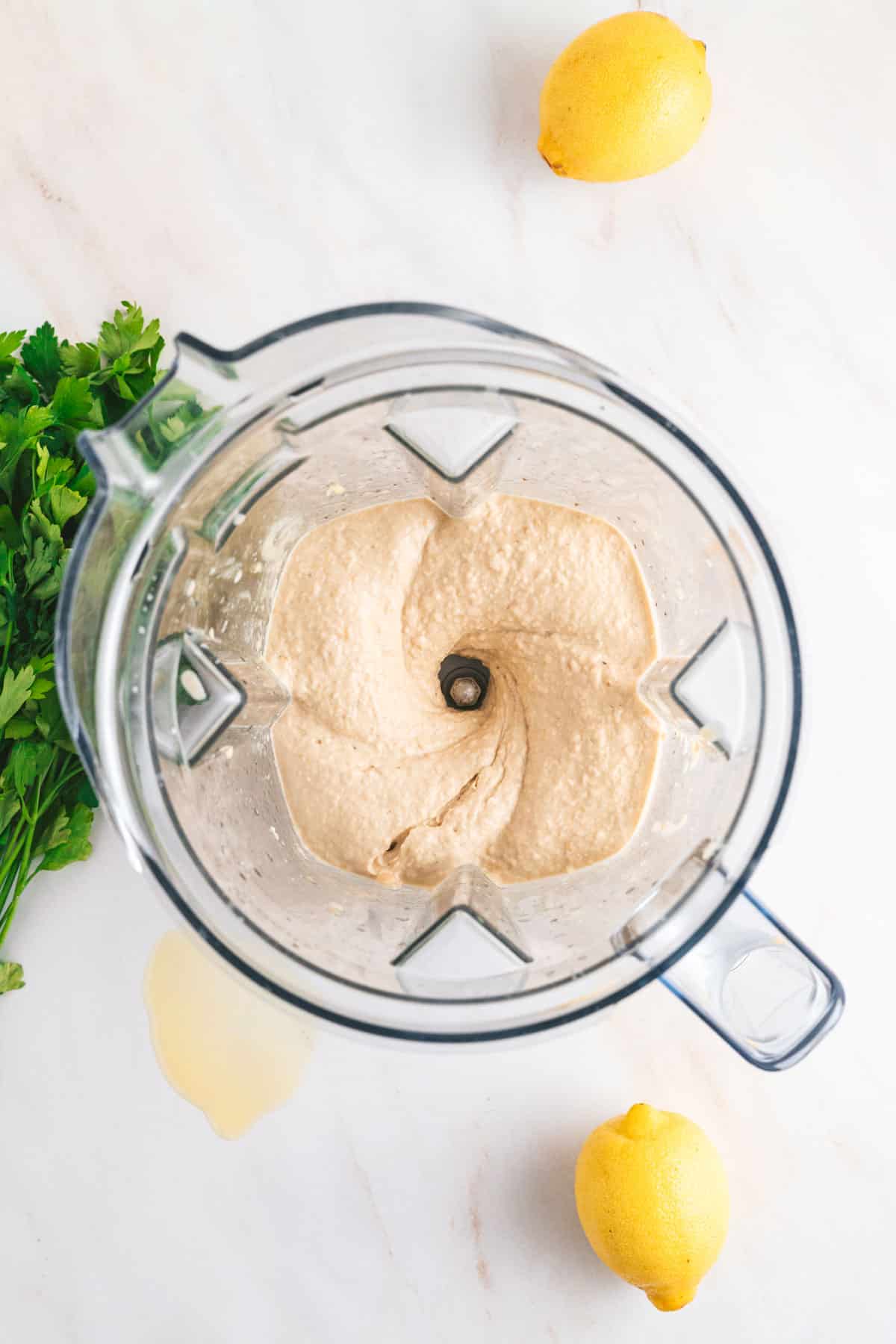 Blender with creamy hummus inside, surrounded by two lemons and parsley on a white surface.