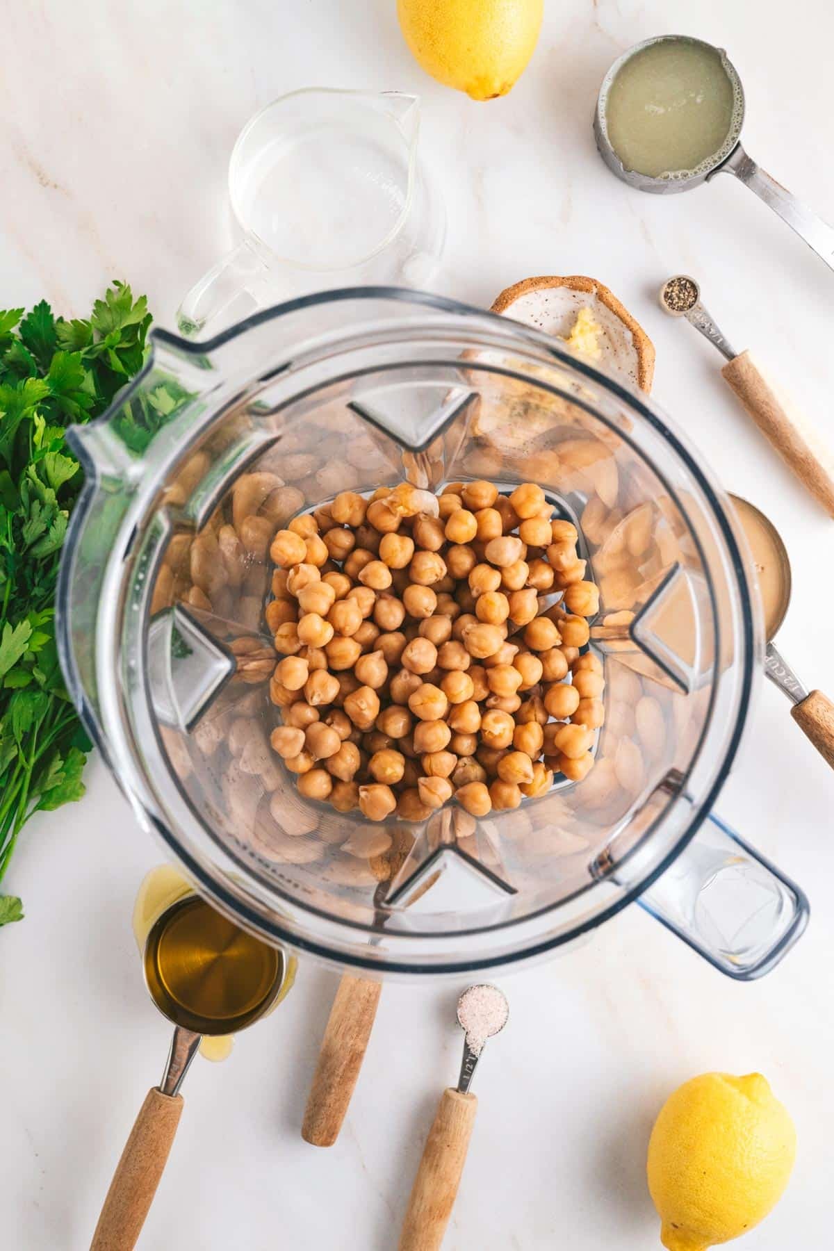 Top view of a blender with chickpeas surrounded by parsley, lemons, measuring spoons with spices, olive oil, and tahini on a kitchen counter.