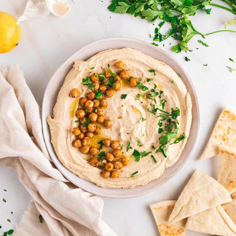 A bowl of hummus garnished with roasted chickpeas and parsley, surrounded by pita bread, fresh parsley, a lemon, and a beige cloth on a white surface.