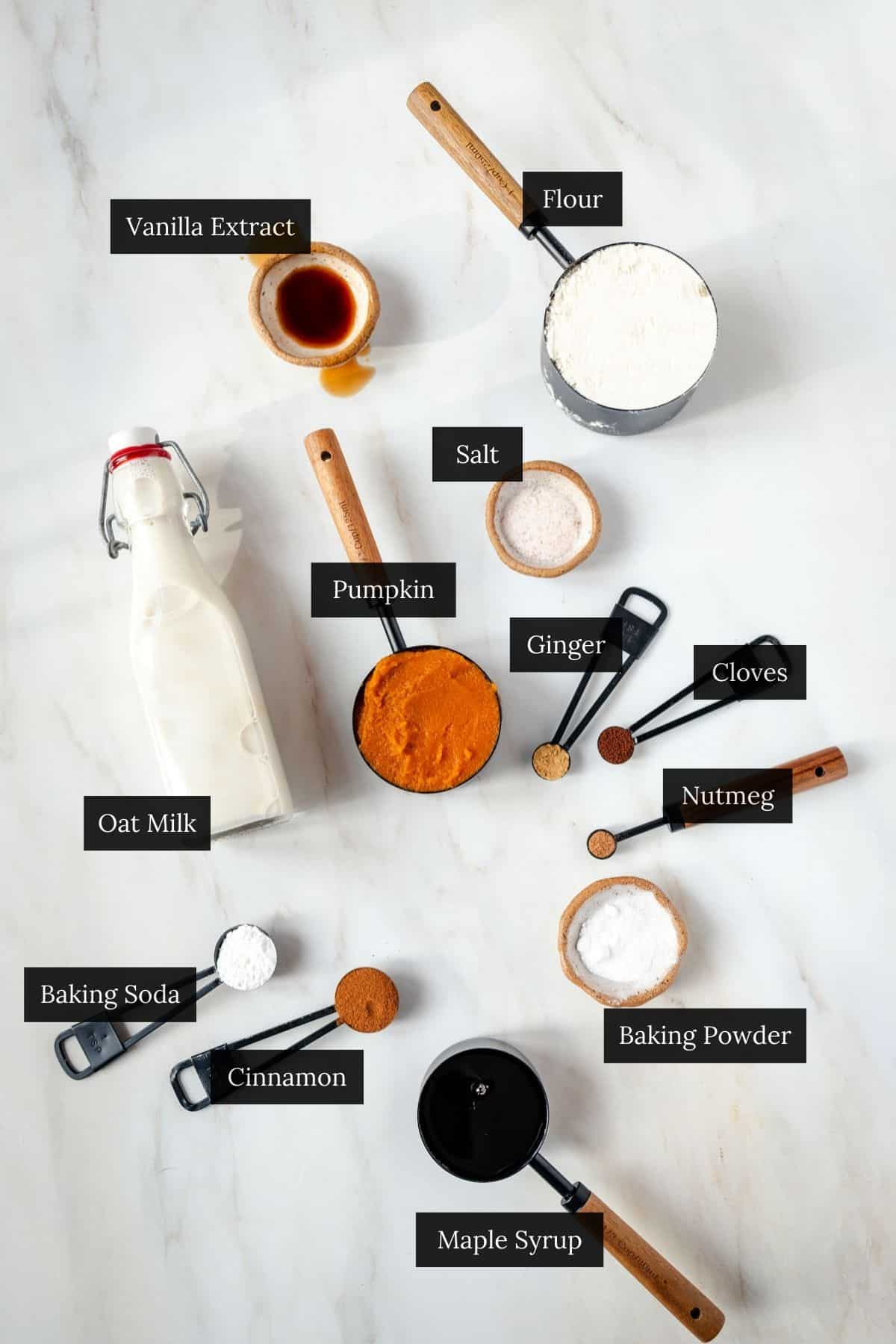 Flat lay of labeled baking ingredients on a white surface, perfect for pumpkin spice donuts, including flour, pumpkin, spices, oat milk, and vanilla extract.