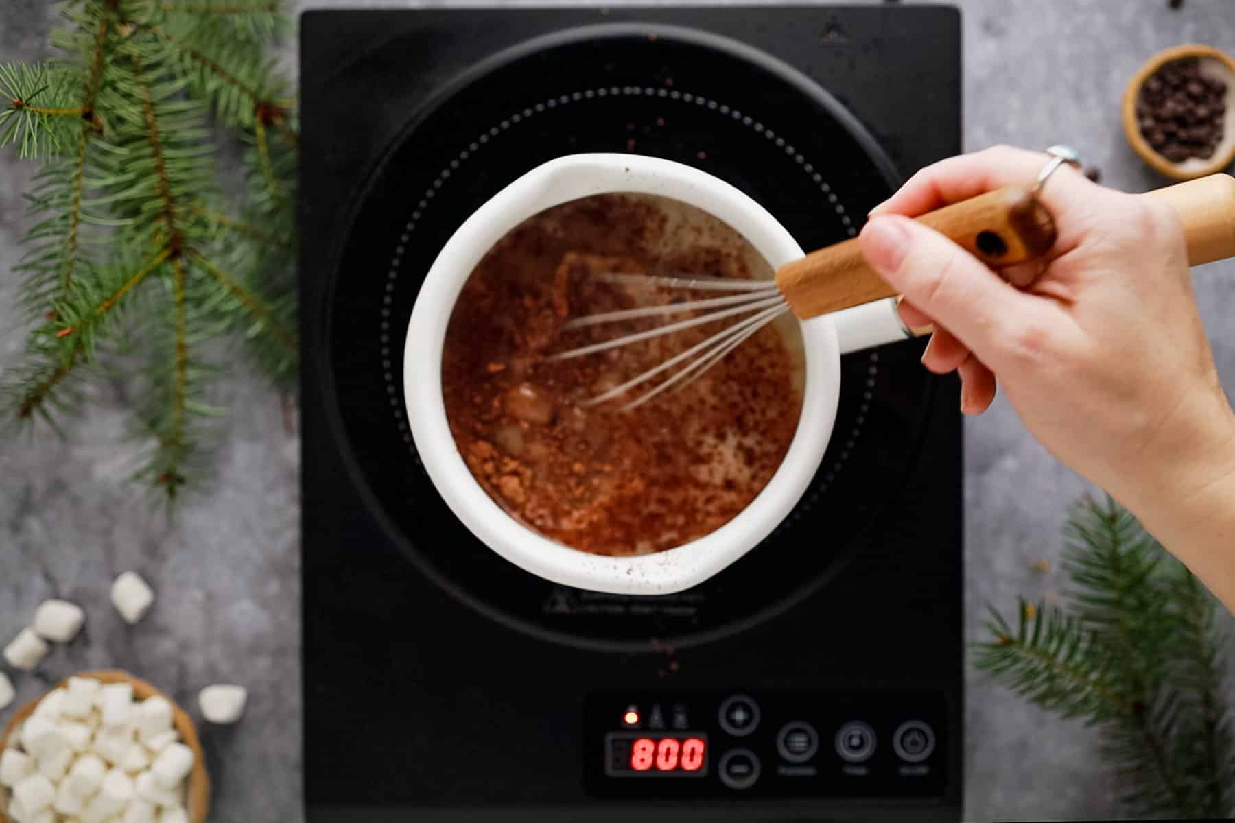 Top view of a person whisking cocoa in a pot on an electric stove, surrounded by pine branches and scattered marshmallows.