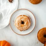 A glazed donut sits on a white plate surrounded by pumpkin spice donuts, with a scattering of cinnamon adorning the marble surface.