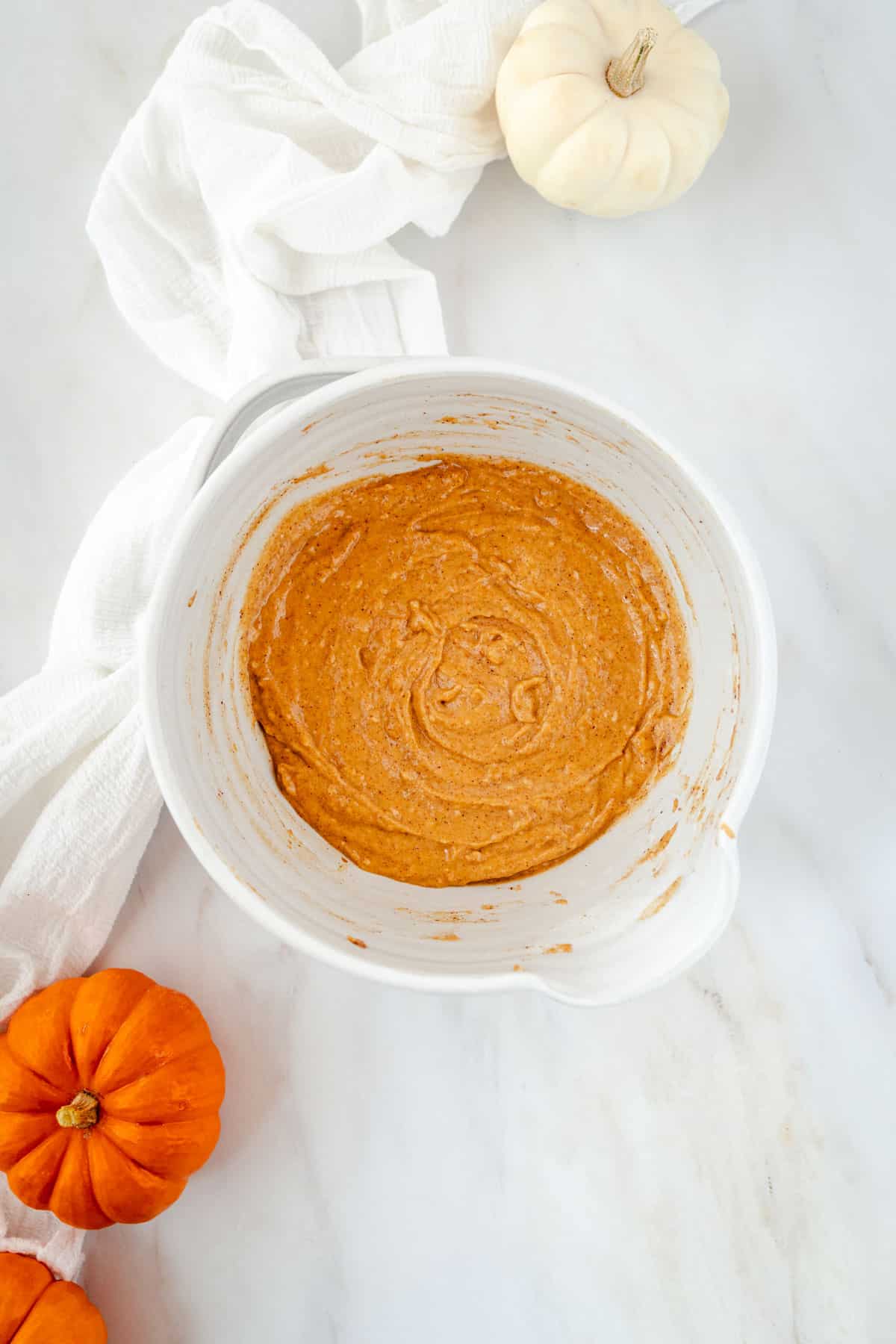 A mixing bowl filled with pumpkin batter on a marble surface, surrounded by small pumpkins and a white cloth.