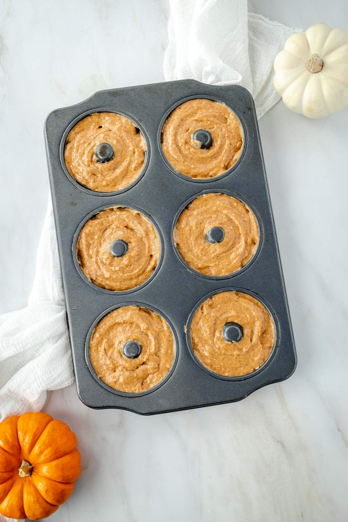 Donut baking pan filled with pumpkin batter next to a small orange pumpkin and a white pumpkin on a light surface.