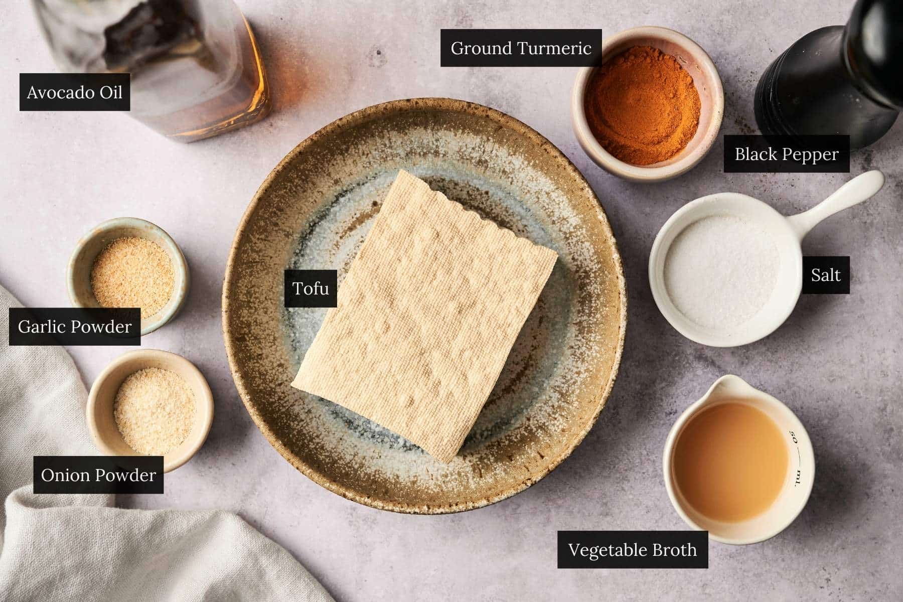A plate with tofu is surrounded by bowls of ground turmeric, black pepper, salt, garlic powder, onion powder, vegetable broth, and a bottle of avocado oil on a light surface.