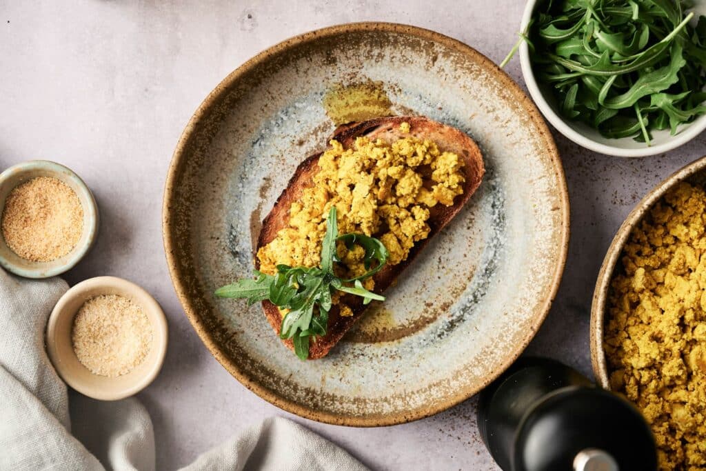 A plate with toast topped with scrambled tofu and arugula. Nearby are bowls of arugula and seasoning, all on a light gray surface.