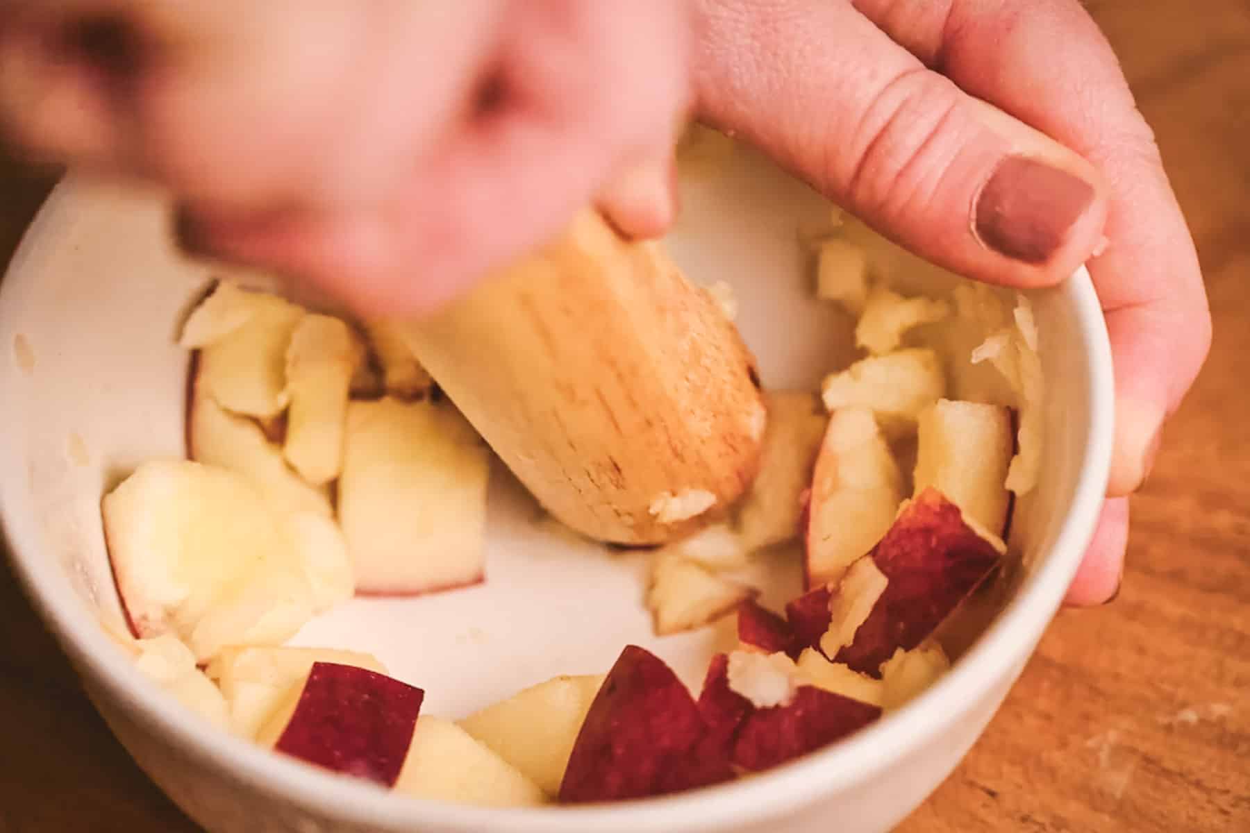 Person muddling apples in a white bowl.