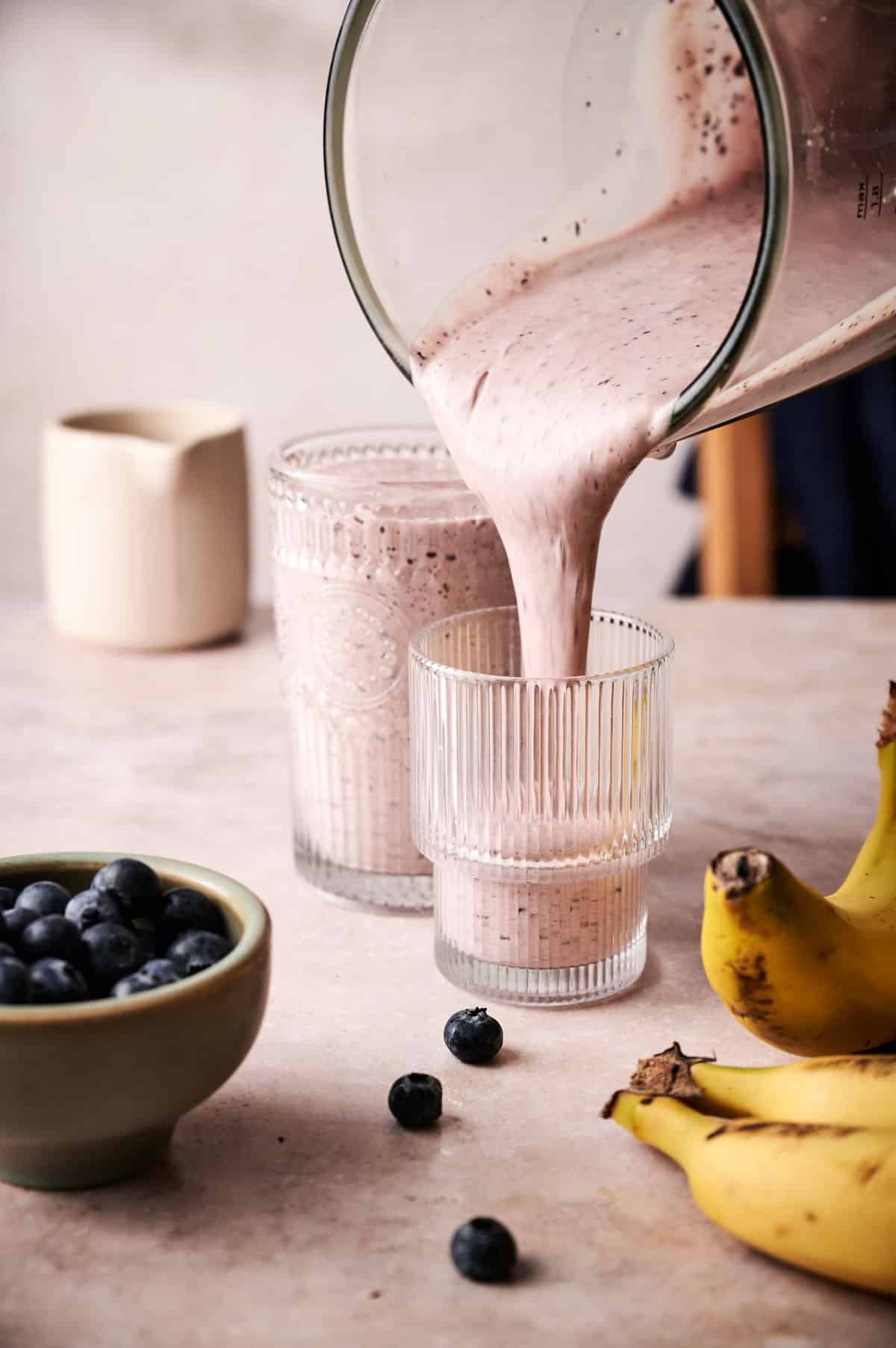 Pouring a raspberry blueberry banana smoothie into clear glass cups.