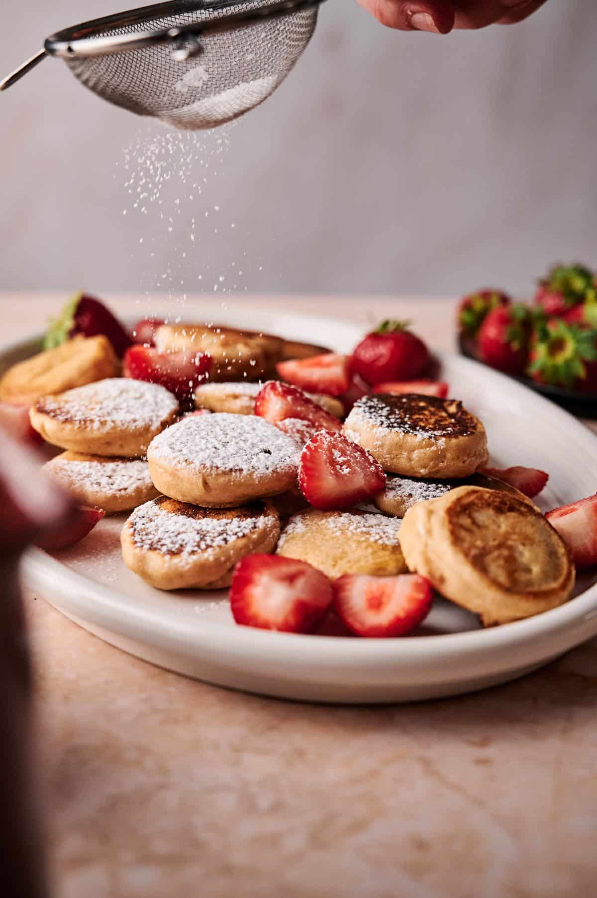 Shaking powdered sugar on top of mini pancakes on an oval plate.