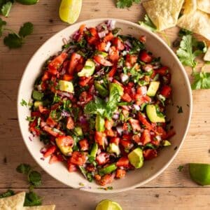 Homemade avocado pico in a shallow cream bowl with lime wedges and cilantro sprigs next to it.