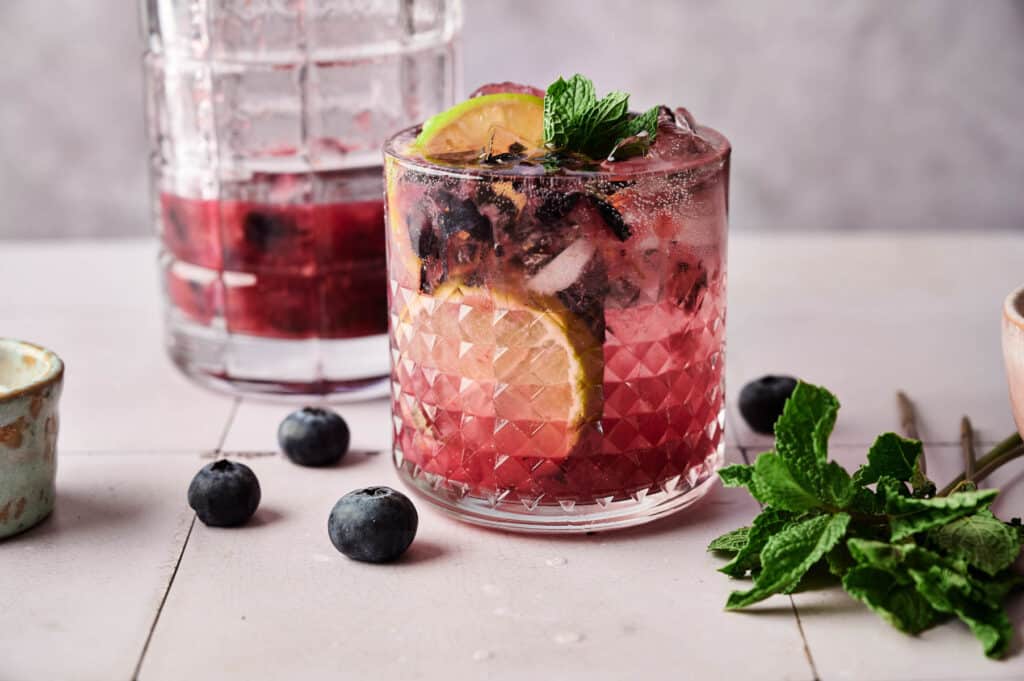 Fresh blueberry smash mocktail in a cocktail glass with lime slices, blueberries andmint in the glass next to a pitcher.