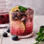 Fresh blueberry smash mocktail in a cocktail glass with lime slices, blueberries andmint in the glass next to a pitcher.