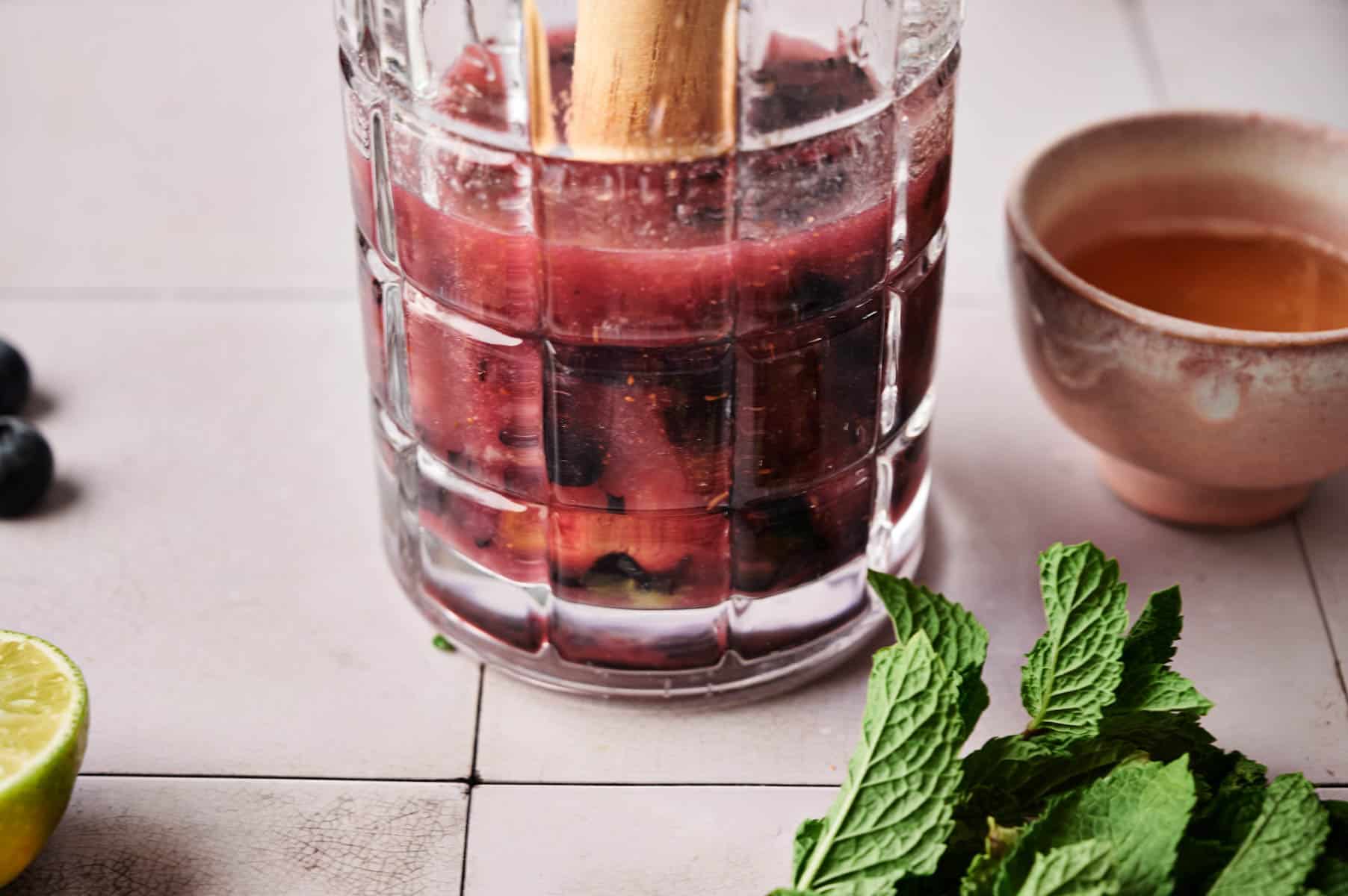 Mashing blueberries with a wooden muddle in a clear glass pitcher.