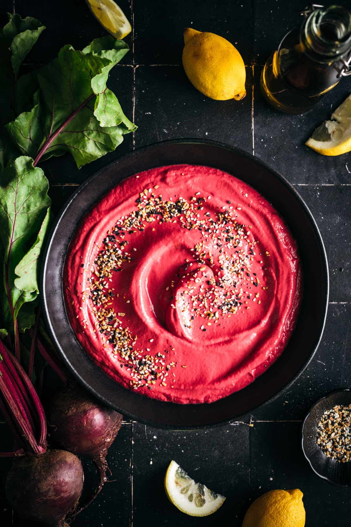 Fresh beet hummus in a low rim black ceramic bowl with fresh beets and lemons next to it and seeds and spices sprinkled on top.