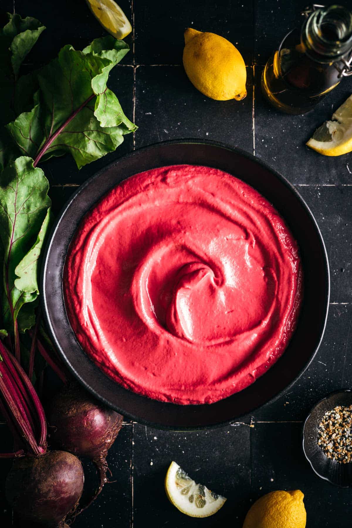 Fresh beet hummus in a low rim black ceramic bowl with fresh beets and lemons next to it.