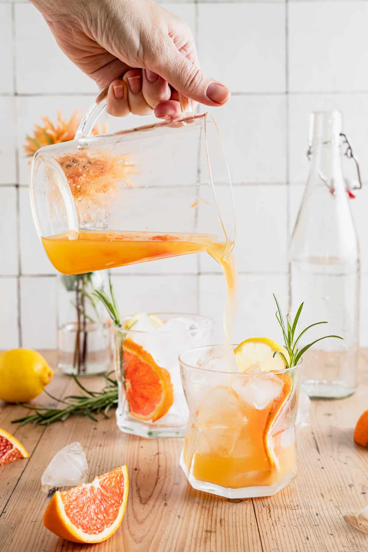 Pouring non-alcoholic citrus mocktail into glass cups with slices of orange, lemon, and fresh rosemary.