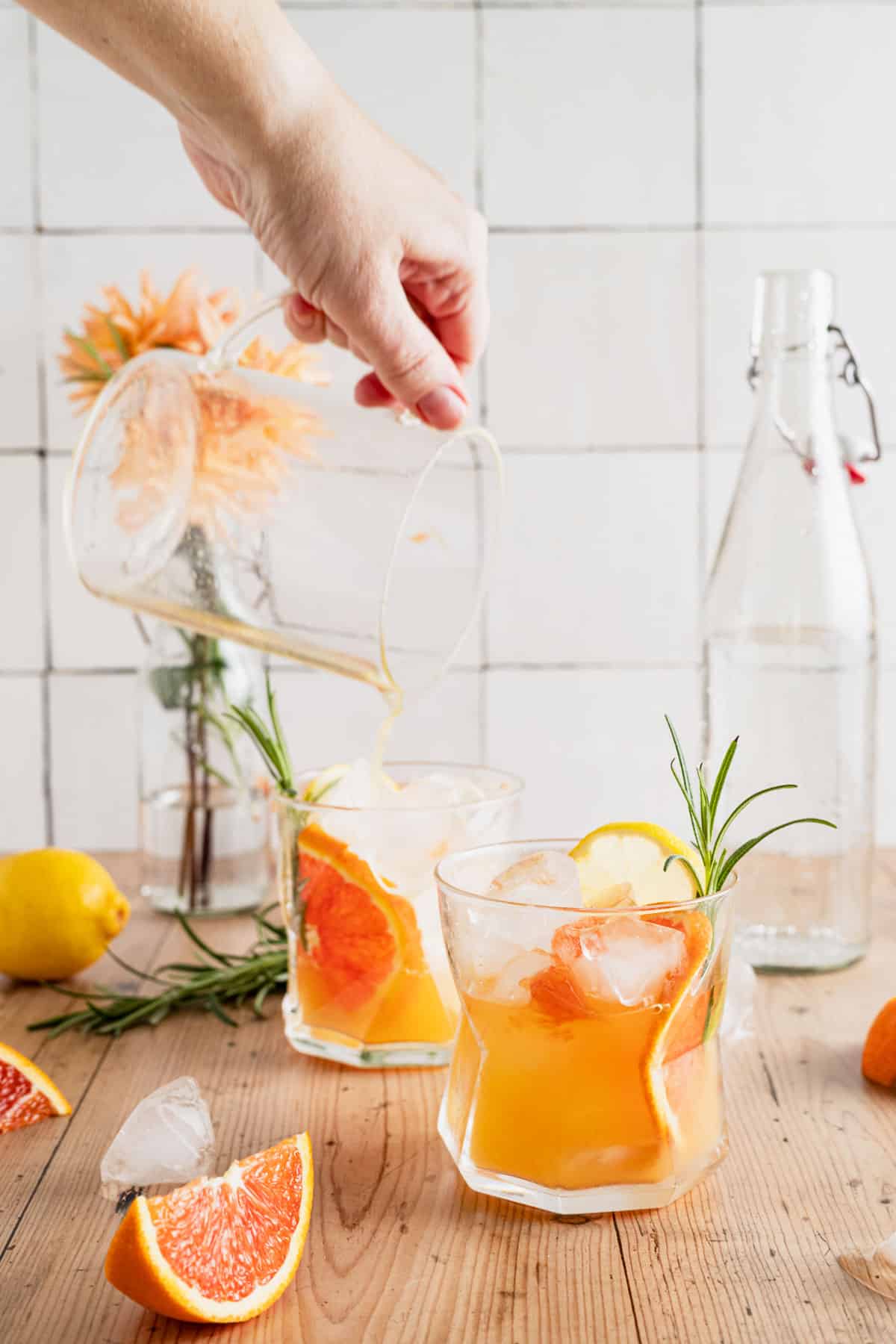 Pouring non-alcoholic citrus mocktail into glass cups with slices of orange, lemon, and fresh rosemary.