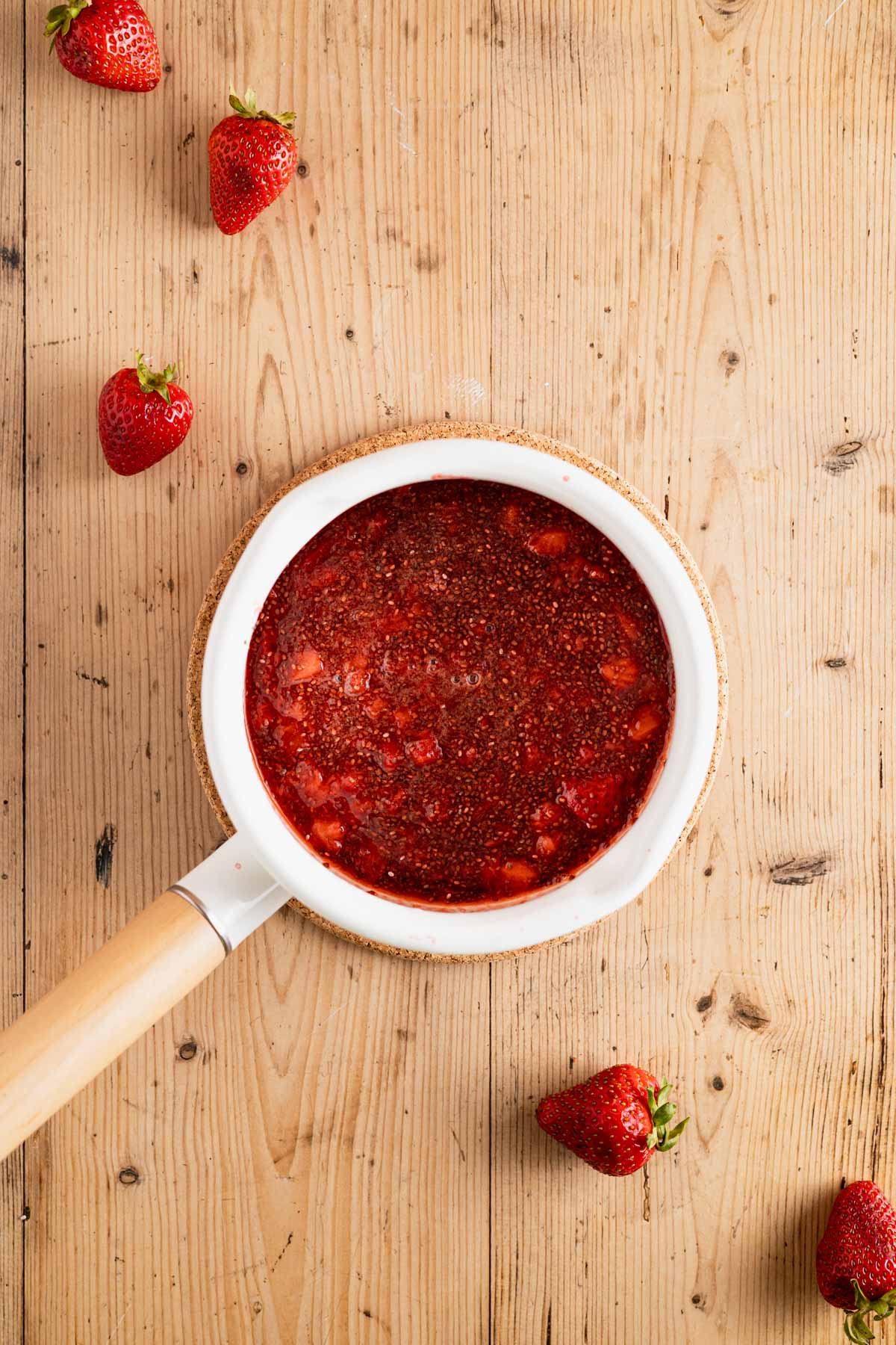 All the ingredients for chia seed strawberry jam in a white saucepan with a wooden handle.
