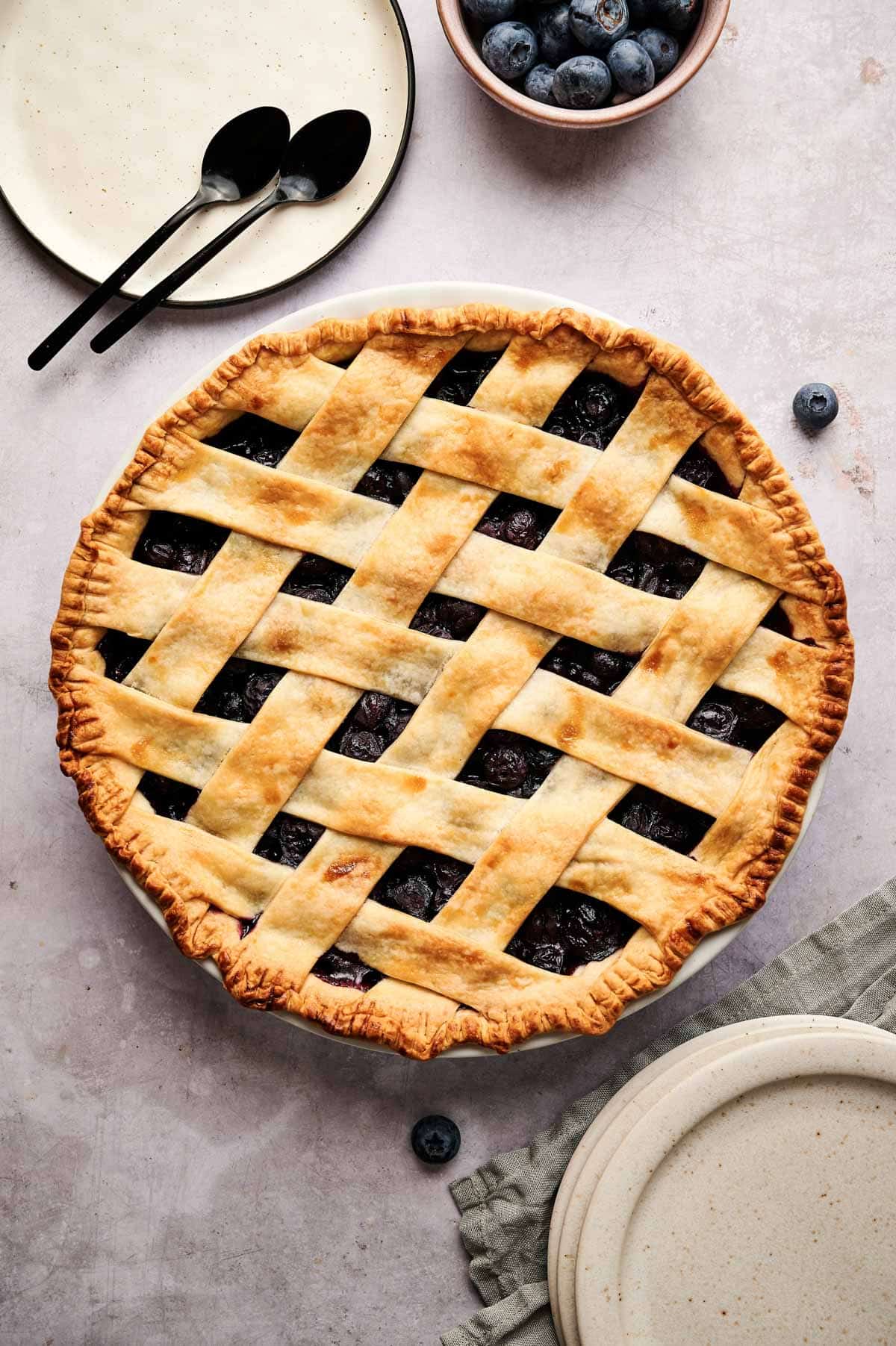 Whole vegan blueberry pie baked golden brown with cream colored plates next to it.