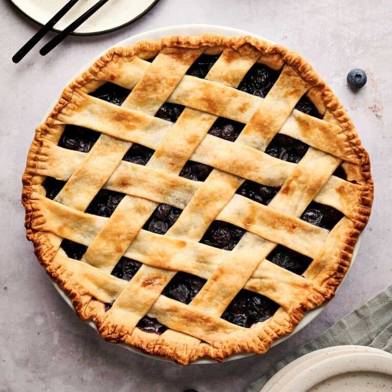 Whole vegan blueberry pie baked golden brown with cream colored plates next to it.