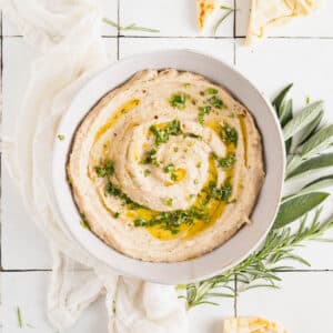 Homemade white bean dip in a white bowl with olive oil drizzled on top and fresh herbs.