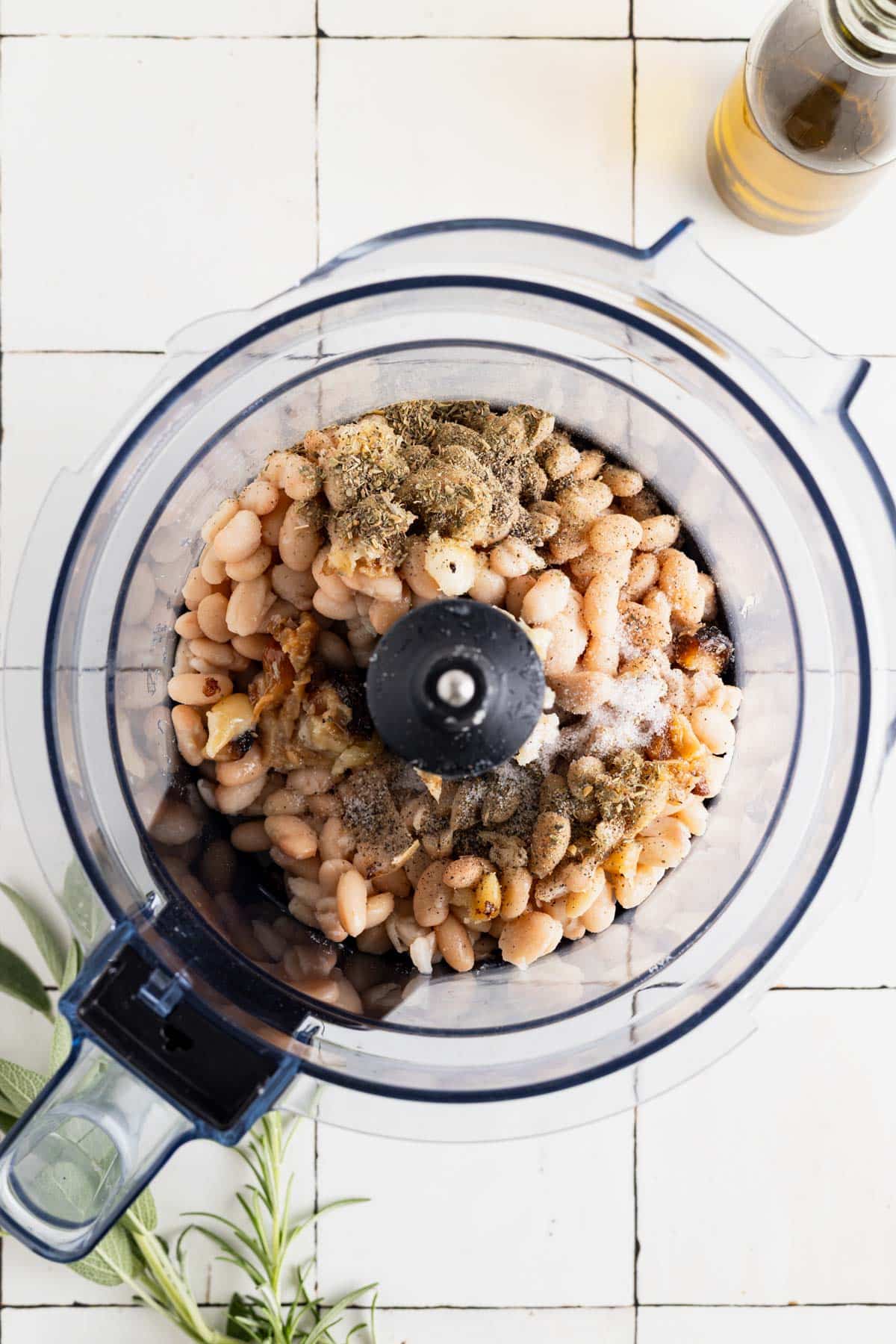 White beans, garlic, herbs, and salt in a food processor on a white tile table.