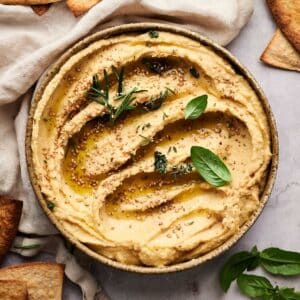 Hummus in a tan shallow bowl with toasted pita and fresh herbs next to it.