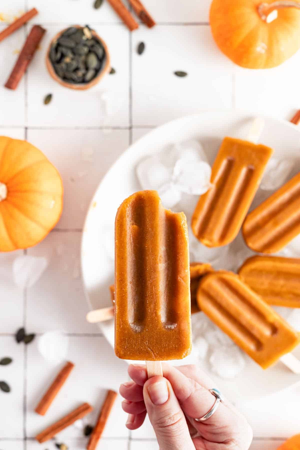 Woman holding vegan pumpkin Popsicle over a white plate with ice cubes.