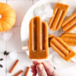 Woman holding vegan pumpkin popsicles over a white plate with ice cubes.