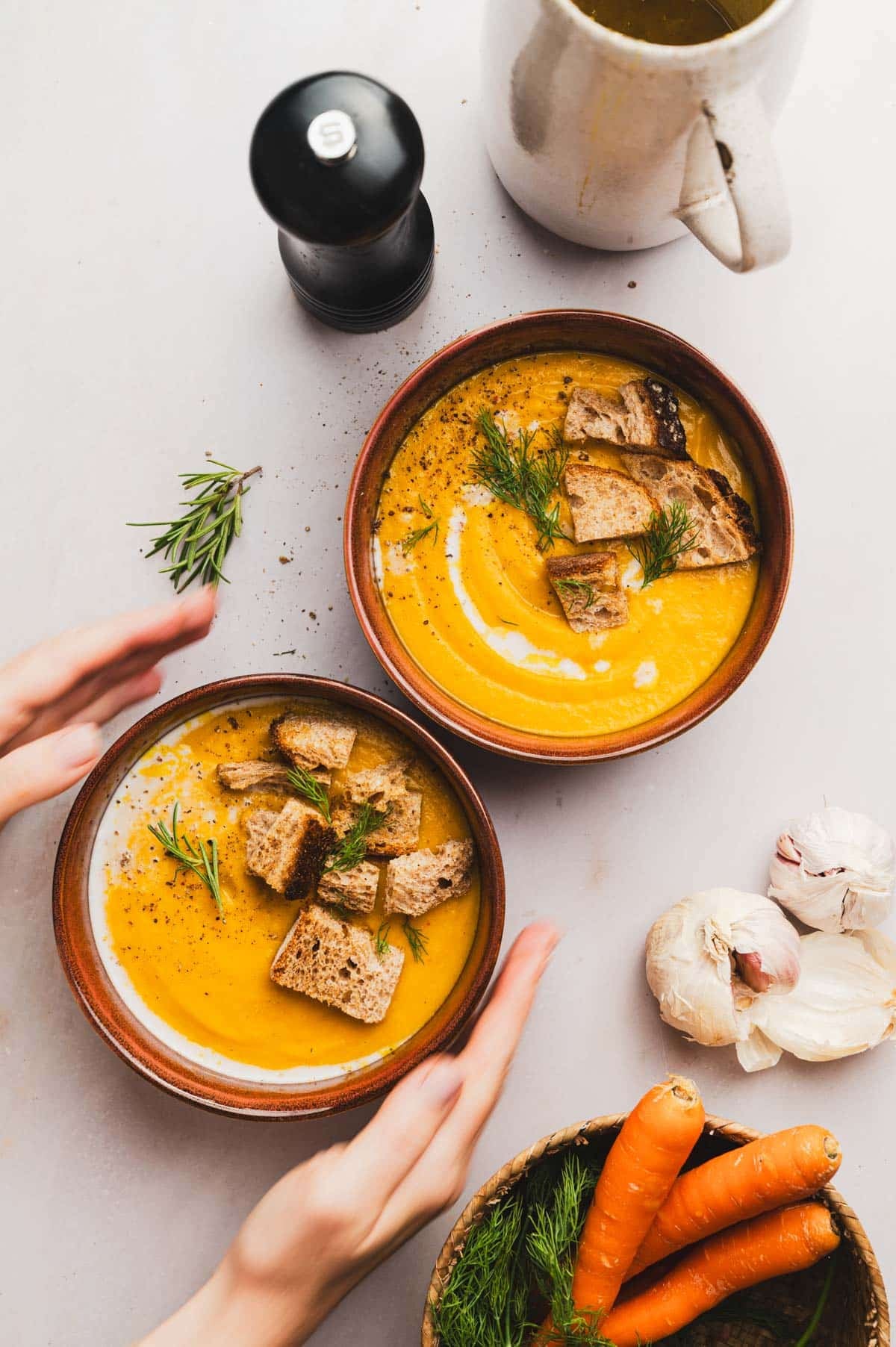 Two bowls of lentil carrot soup on a counter with fresh carrots and garlic next to it.