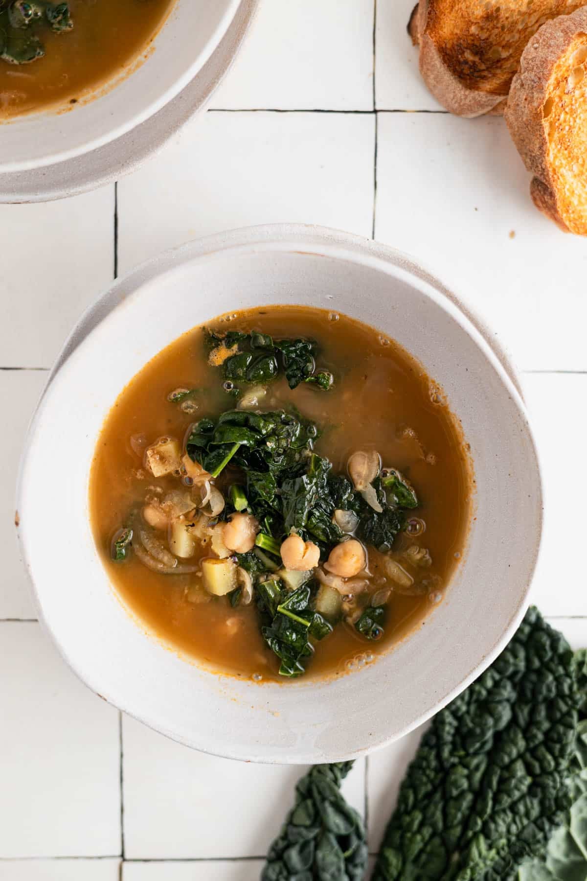 White ceramic bowl filled with chickpea soup with crispy bread and fresh kale next to it.