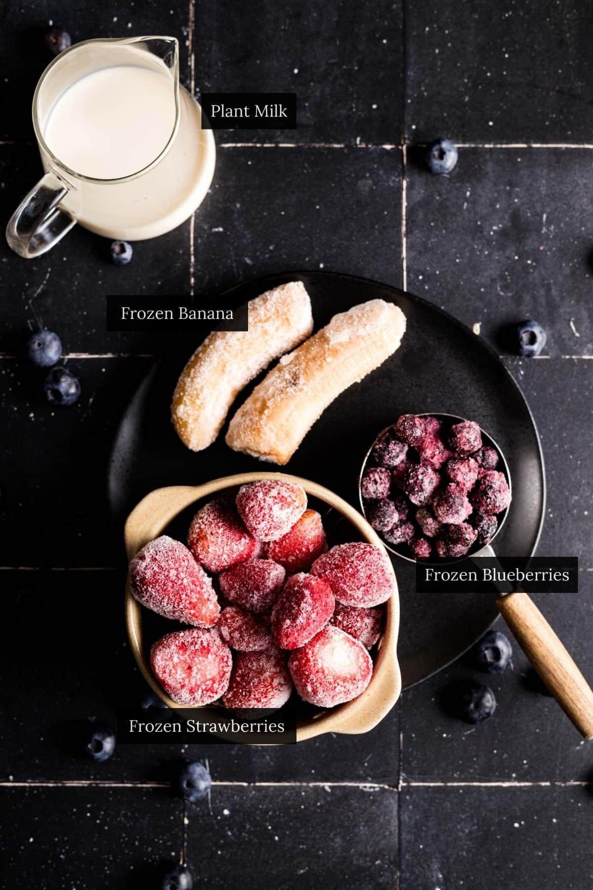 Ingredients for a strawberry banana and blueberry smoothie on a black tiled counter.